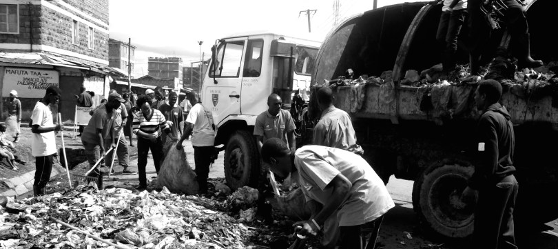 Men and women rake and shovel a plie of garbage into bags as part of the Kariobangi Waste Management Allianc
