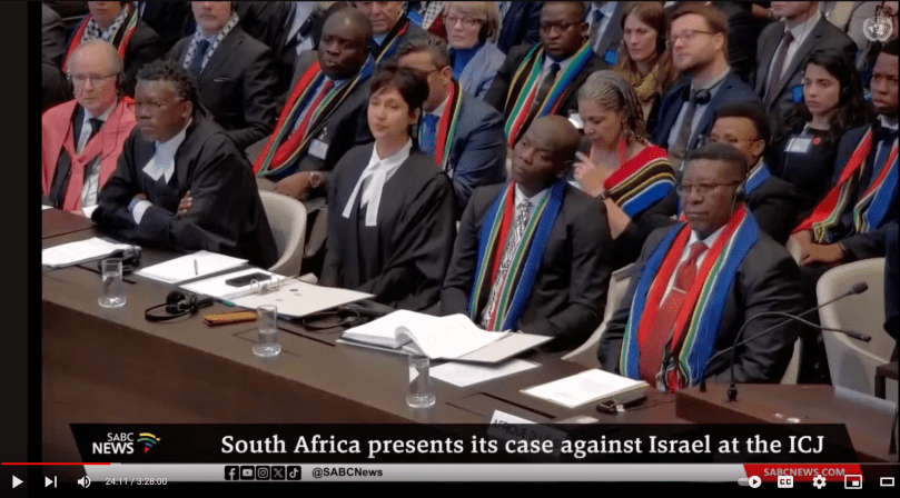 Five formally dressed lawyers sit at a long brown wood table at the International Court of Justice. They are South Africa's delegation, each dressed in black suits: three men have dark African skin, one lighter skin woman in the middle, and one European-descent man on the left. Two of them, and many people seated behind them wear long cloths with the colors of the post-Apartheid South African flag. A South African Broadcast Corporation news chyron at the bottom reads: "South Africa presents its case against Israel at the ICJ"