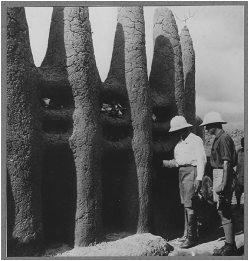 Vue de Marcel Griaule et de Michel Leiris posant devant le sanctuaire du kono du quartier de la chefferie de Kéméni. Construit en banco, à la fois matériau et technique , le sanctuaire comporte des niches contenant des ossements d’animaux sacrifiés. (Catalogue MQBJC)